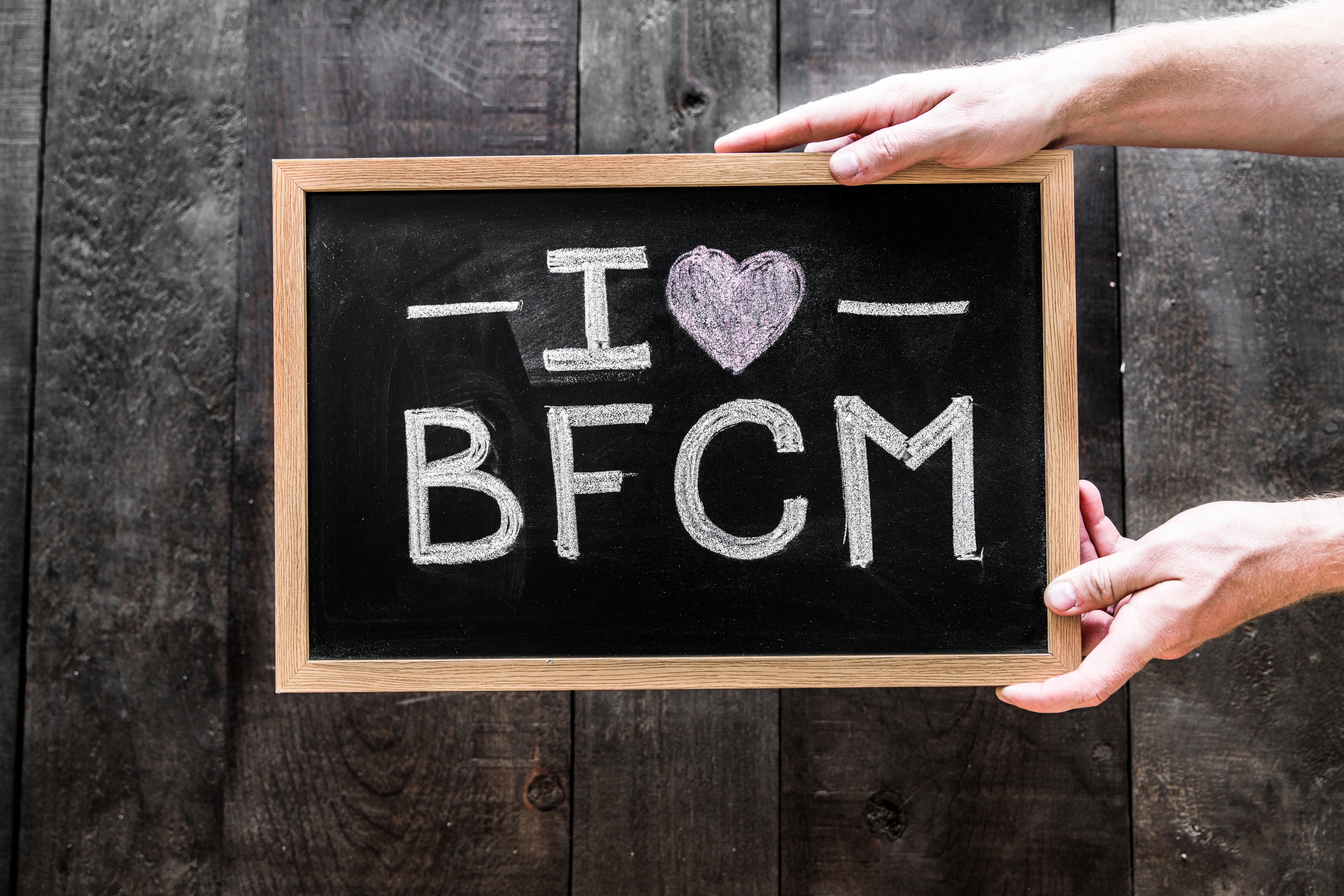 I heart BFCM on a black board being held in front of a wooden background. Black Friday/Cyber Monday Promotion